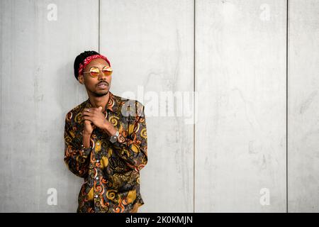 Nachdenklicher afroamerikanischer Kerl mit Dreadlocks, der eine stilvolle runde Sonnenbrille mit rotem Stirnband und einem farbenfrohen Hemd trägt, das wegschaut Stockfoto