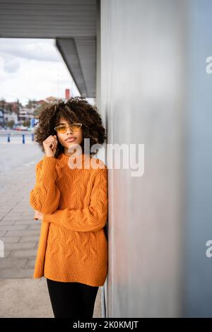 Schöne ethnische Frau mit lockigen Afro-Haaren trägt einen leuchtend orangefarbenen Pullover und blickt auf die Kamera gegen die graue Wand Stockfoto