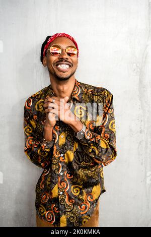 Fröhlicher afroamerikanischer Kerl mit Dreadlocks, der eine stilvolle runde Sonnenbrille mit rotem Stirnband und einem farbenfrohen Hemd trägt, das lächelt und die Kamera anschaut Stockfoto