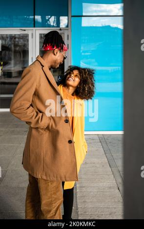 Fröhlicher afroamerikanischer Mann und Frau in stilvollen beige- und gelben Mänteln umarmen sich gegenseitig, während sie auf der Straße gehen und einander angucken Stockfoto