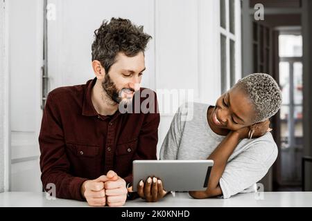 Zufriedene afroamerikanische Frau und bärtiger Mann lächeln, während sie am Tisch sitzen und zusammen ein digitales Tablet zu Hause anschauen Stockfoto