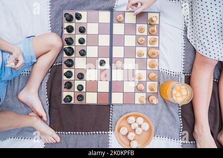 Barfuß beschnittenes Mädchen mit Bruder, der Schach spielt, während er auf Textil mit einem Krug mit Getränk und Keksen auf dem Rasen sitzt Stockfoto