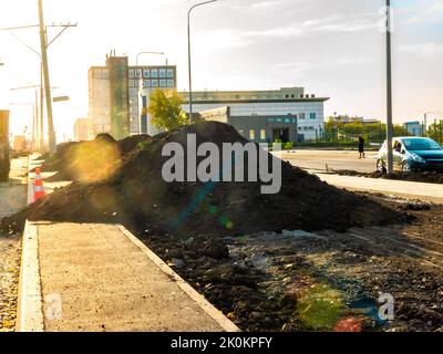 Auf der Fahrbahn und teilweise auf dem Bürgersteig stapeln sich Erdhaufen, um einen Rasen oder einen grünen Streifen zu arrangieren, selektiver Fokus und Hintergrundbeleuchtung Stockfoto