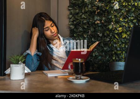 Fokussierte junge hispanische Universitätsstudentin, die über Kopfhörer Musik hört und Buch liest, während sie mit einer Tasse Kaffee und einem Glas Kaffee am Tisch sitzt Stockfoto