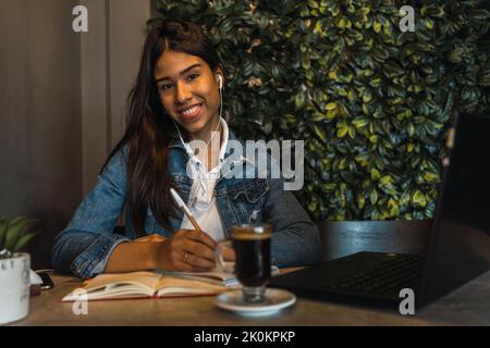 Fröhliche junge hispanische Frau mit Ohrhörern, die die Kamera betrachten, während sie online studiert und Videovorträge auf dem Laptop ansieht und sich Notizen mit Copyboo ansieht Stockfoto