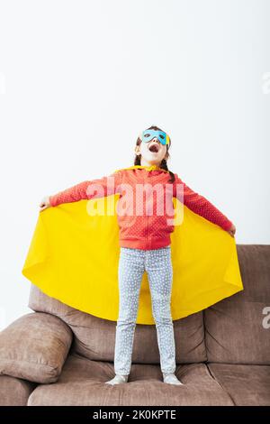 Voller Körper von selbstbewussten Kind in Superhelden Umhang und Maske auf dem Sofa stehend mit dem Mantel offen vorgetäuscht, in hellen Raum mit weißer Wand fliegen Stockfoto