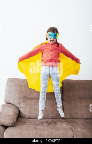 Voller Körper von selbstbewussten Kind in Superhelden Umhang und Maske springen auf dem Sofa mit dem Mantel offen vorgibt, in hellen Raum mit weißer Wand fliegen Stockfoto