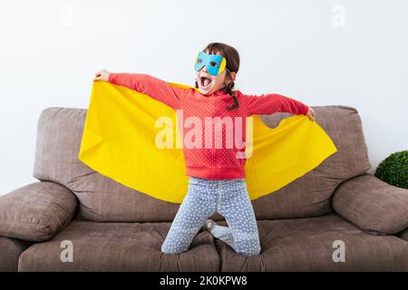 Selbstbewusstes Kind im Superhelden-Umhang und Maske auf dem Sofa mit offenem Mantel, der vorgibt, im hellen Raum mit weißer Wand zu fliegen Stockfoto