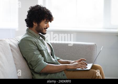 Seitenansicht eines indischen Mannes, der auf der Couch saß und einen Laptop benutzte Stockfoto