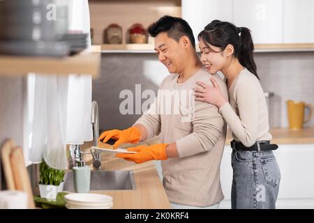 Glückliche asiatische Frau unterstützt ihren Mann Geschirr waschen Stockfoto