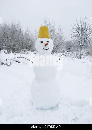 Ein Schneemann aus drei Schneebällen, er hat einen gelben Eimer auf dem Kopf, eine Karotte statt einer Nase und Kohlen statt Augen. Vor dem Hintergrund von Stockfoto