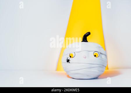 Kreative Halloween Hintergrund. Niedliche und schüchterne Kürbis-Mumie mit Spinne im gelben Projektor-Lichtstrahl auf dem weißen Hintergrund. Kürbis mit verwirrt lo Stockfoto