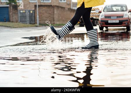 Frau, die nach dem Regen auf der Straße Spaß hat. Nahaufnahme weiblicher Beine in Regengummistiefeln, die in die Pfütze mit Wasserspritzern und Tropfen in der Lig bei Sonnenuntergang gehen Stockfoto