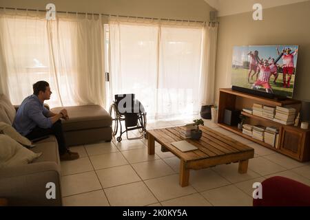 Mann mit einer Behinderung sitzt auf der Couch und schaut sich ein Fußballspiel an Stockfoto