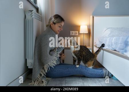Junge Frau mit warmer Decke bedeckt spielt mit Katze fütterndes Tier mit Essen auf dem Boden sitzen Stockfoto