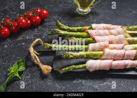 Frischer grüner Spargel, eingewickelt in Speck auf einem schwarzen Steintisch Stockfoto