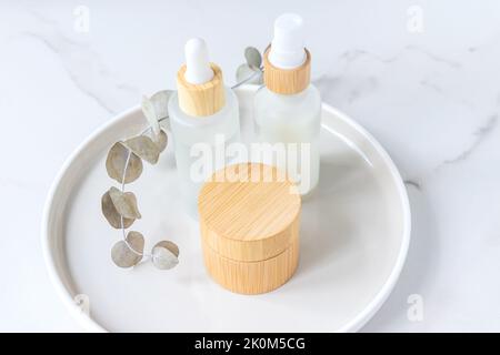 Glas aus Bio-Creme, Gesichts-Serum in einer Tropfflasche mit Eukalyptusblättern auf einem Tablett auf Marmor. Make-up-Kosmetikset ohne Markenaufdruck. Natürliches Bio-Produkt Stockfoto