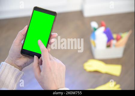 Frau mit Smartphone mit Chroma-Bildschirm. Smartphone mit einem grünen Bildschirm auf dem Hintergrund von Waschmitteln. Stockfoto