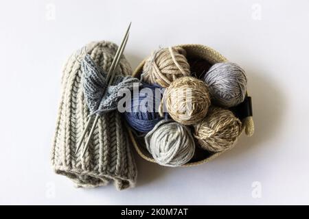 Graue, braune und blaue Strickgarnkugeln in einem beigen Korb mit Stricknadeln und Strickmützen für Winter und Herbst, Strickhobby Stockfoto