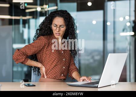 Büroangestellter in Lateinamerika ist am Arbeitsplatz krank, eine übermüdete Geschäftsfrau hat Rückenschmerzen und massiert ihren Rücken mit der Hand. Stockfoto