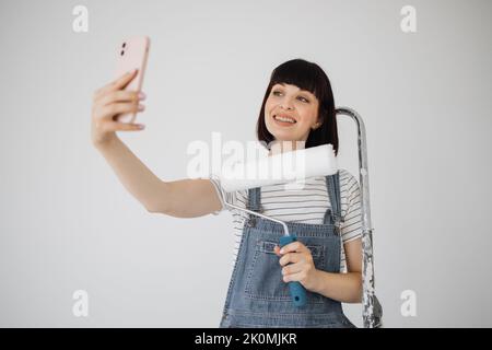 Rote Haare Mädchen mit niedlichem Lächeln hält eine Malrolle und Smartphone in den Händen. Junge Frau, die sich auf eine Leiter stützt, die sie bei Reparaturen verwendet, und Videoanruf am Telefon hat. Stockfoto