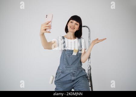 Rote Haare Mädchen mit niedlichem Lächeln hält eine Malrolle und Smartphone in den Händen. Junge Frau, die sich auf eine Leiter stützt, die sie bei Reparaturen verwendet, und Videoanruf am Telefon hat. Stockfoto