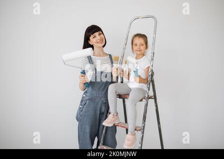 Eine fröhliche glückliche Familie renoviert eine erworbene Wohnung. Die Mutter umarmt ihre Tochter, sitzt auf einer Leiter, mit Freude. Jeder hält eine Farbrolle und Pinsel, um die Wände zu malen. Stockfoto