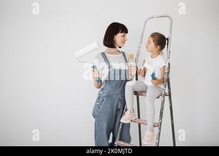 Eine fröhliche glückliche Familie renoviert eine erworbene Wohnung. Die Mutter umarmt ihre Tochter, sitzt auf einer Leiter, mit Freude. Jeder hält eine Farbrolle und Pinsel, um die Wände zu malen. Stockfoto