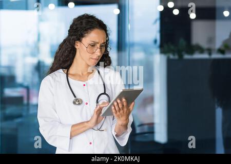 Ernsthaft denkende hispanische Ärztin mit Tablet-Computer, Ärztin in Brille und weißer medizinischer Mantel mit Stethoskop, die in der Büroklinik arbeitet. Stockfoto