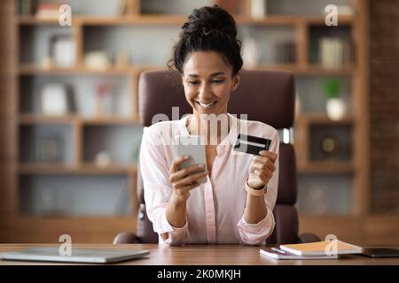 Positive Geschäftsfrau, die online im Büro mit dem Smartphone einkauft Stockfoto