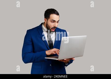 Überraschender bärtiger Mann mit Laptop, Tastatur, Surfen im Internet, Freiberufler, der am Computer arbeitet und einen offiziellen Anzug trägt. Innenaufnahme des Studios isoliert auf grauem Hintergrund. Stockfoto