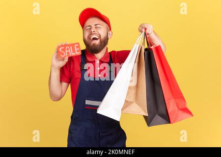 Porträt eines fröhlichen, lustigen Kuriermanns, der mit Einkaufstaschen und einer Karte mit Verkaufsaufschrift steht, lacht, blaue Overalls und Mütze trägt. Innenaufnahme des Studios isoliert auf gelbem Hintergrund. Stockfoto