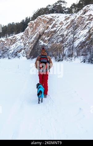 Rückansicht junger Mann in rotbrauner warmer Kleidung mit Rucksack mit Mischlingshund in warmem blauem Anzug, der im Winter AC zwischen Felsen und Klippen im Schnee läuft Stockfoto