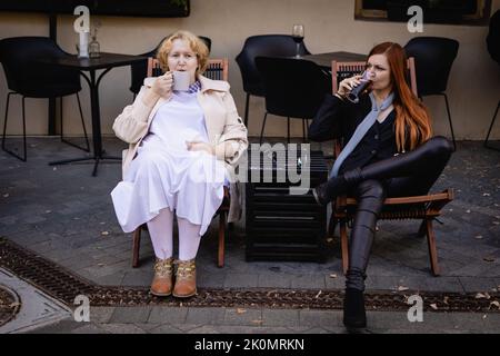 Zwei Frauen zusammen - blond in hellen und rothaarig in dunklen Kleidern trinken Kaffee und Wein, sitzen an einem Tisch eines Straßencafés. Stockfoto