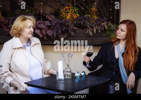 Zwei Frauen zusammen - blonde in hellen und rothaarige in dunklen Kleidern, die an der Bar eines Straßencafés reden. Stockfoto