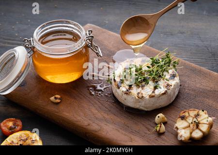 Der Camembert-Käse ist rund, gegrillt, mit goldenem Honig auf einem hölzernen Schneidebrett gegossen. Weicher selektiver Fokus, schwarzer Hintergrund.Platz zum Kopieren. Gesunde Ernährung. Stockfoto