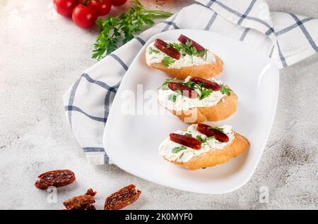 CANape oder Crostini mit geröstetem Baguette, Frischkäse, sonnengetrockneten Tomaten, Kräutern auf weißem Teller. Handgemachte leckere Sandwiches zum Frühstück. Stockfoto