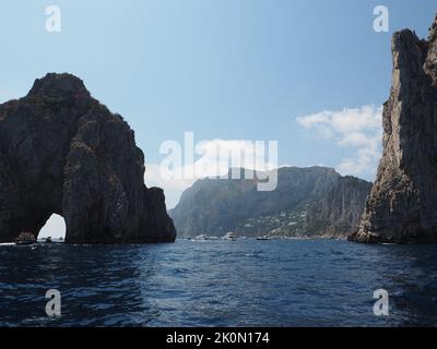 Die Küste der Insel Capri ist sehr felsig und steil. Dies sind die ikonischen Faraglioni Felsen, Symbole der Insel. Touristenboote fahren durch den Bogen. Stockfoto