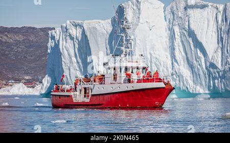 Leuchtendes rotes Fischerschiff, das am 17. Juli 2022 Touristen vor dem aufragenden Eisberg im Ilulissat-Eisfjord in Grönland transportiert Stockfoto