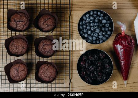 Frisch gebackene Schokoladen-Cupcakes mit Zutaten in der Nähe. Garnieren Sie mit frischen Heidelbeeren und Brombeeren und Marmelade Stockfoto