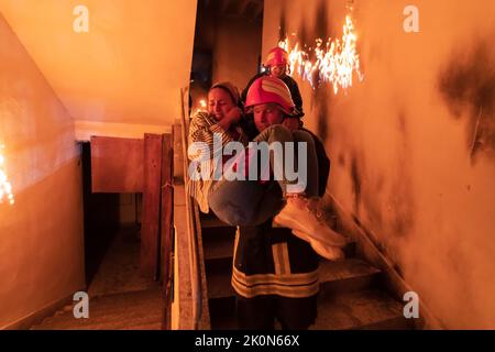 Der tapfere Feuerwehrmann steigt die Treppe eines brennenden Gebäudes hinab und hält das gerettete Mädchen in seinen Armen. Offenes Feuer und ein Feuerwehrmann im Hintergrund. Stockfoto