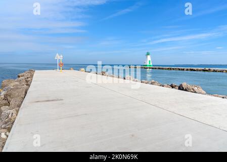 Betonweg auf dem Wellenbrecher am Eingang eines Hafens auf einem See. Auf dem Wellenbrecher auf der anderen Seite des Kanals befindet sich ein Leuchtturm. Stockfoto