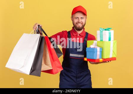 Porträt eines lächelnden, positiv optimistischen, bärtigen Kuriermanns, der mit vielen vorhandenen Schachteln und Einkaufstaschen von Tür zu Tür geliefert wurde. Innenaufnahme des Studios isoliert auf gelbem Hintergrund. Stockfoto