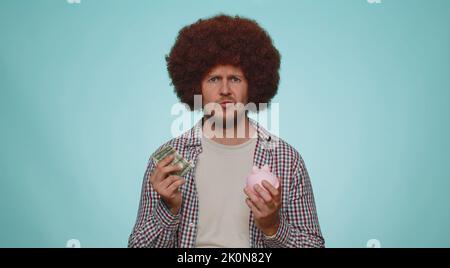 Armer Mann verärgerte sich über unzureichende Geldmenge, hielt ein Sparschwein und eine Dollar-Banknote. Finanzkrise. Insolvenz. Armut und Elend. Junger Mann isoliert auf blauem Studio Wand Hintergrund Stockfoto