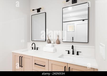 Ein modernes Badezimmer mit einem Schrank aus Holz, schwarzen Wasserhähnen, einer Arbeitsfläche aus weißem Marmor und eckigen Spiegeln mit schwarzen Rändern. Stockfoto