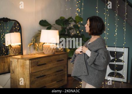 Eine junge Schwangerin sieht sich selbst im Spiegel an. Nahaufnahme Porträt-Foto einer Schwangeren in einem warmen Strickpullover . Konzept der glücklichen Schwangerschaft, Mutterschaft, erwarten Baby. Stockfoto