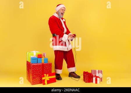 In voller Länge Porträt eines älteren Mannes mit grauem Bart im weihnachtsmann-Kostüm stehend mit Joystick, Videospiel, umgeben von Weihnachtsgeschenken. Innenaufnahme des Studios isoliert auf gelbem Hintergrund Stockfoto