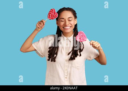 Aufgeregt Frau Frau mit schwarzen Dreadlocks hält zwei Bonbons mit Herzform, hält die Augen geschlossen, hat glücklich positiven Ausdruck, trägt weißes Hemd. Innenaufnahme des Studios isoliert auf blauem Hintergrund. Stockfoto