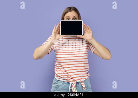 Porträt einer schockiert erstaunten blonden Frau, die ein gestreiftes T-Shirt trägt, das die Hälfte des Gesichts mit einem Tablet bedeckt und ein leeres Display mit Platz für Kopien zeigt. Innenaufnahme des Studios isoliert auf violettem Hintergrund. Stockfoto