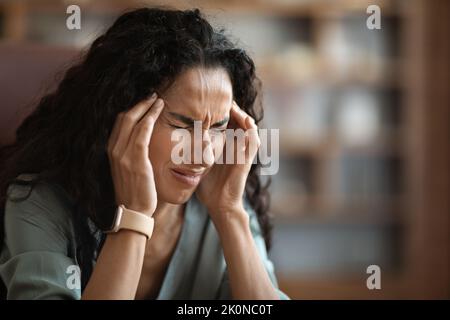 Gestresste Frau, die unter Kopfschmerzen leidet, ihren Kopf berührt, ihr Büroinnenraum Stockfoto